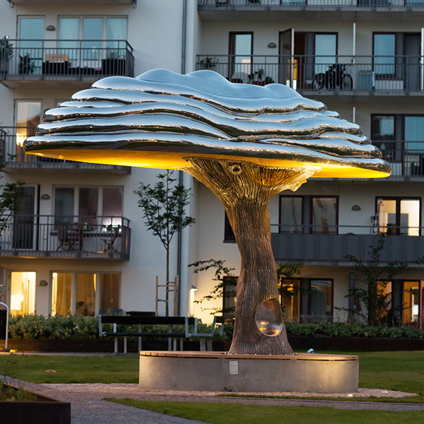 En flera meter hög silvrig skulptur föreställandes ett ekträd. Skulpturen står på en innegård i kvällsljus och är belyst underifrån.