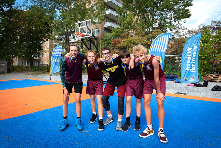 Fem ungdomar i träningskläder poserar med medaljer om halsen på en basketplan i Larsbergsparken på Lidingö.