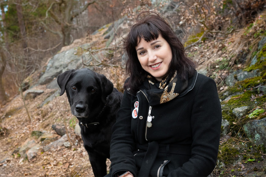 Mörkhårig kvinna med stor svart hund på skogsstig. Båda tittar in i kameran.