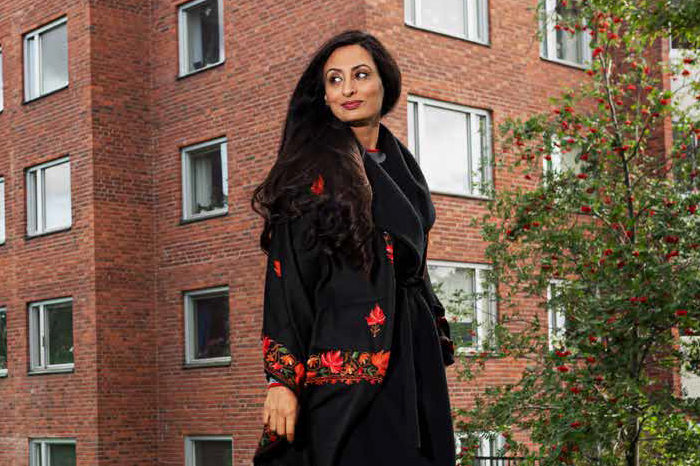 Kvinna i kappa promenerar framför tegelhus och buskage.