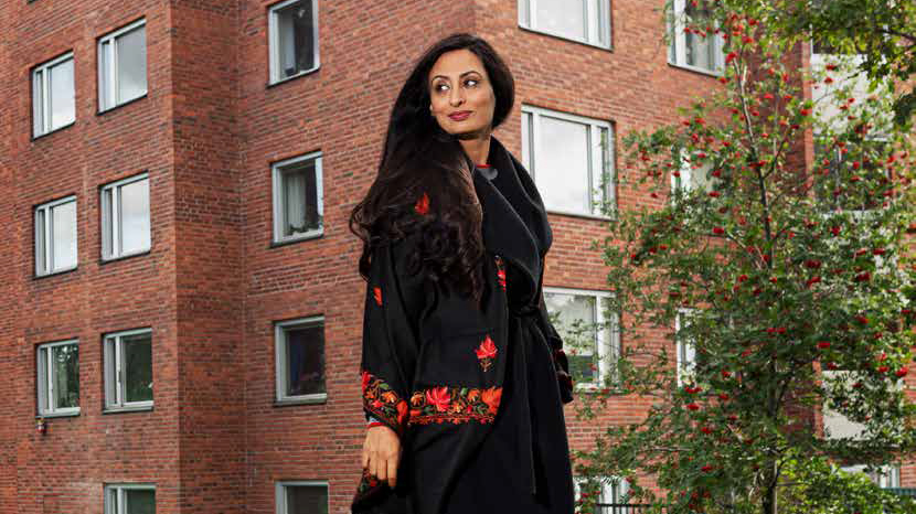 Kvinna i kappa promenerar framför tegelhus och buskage.
