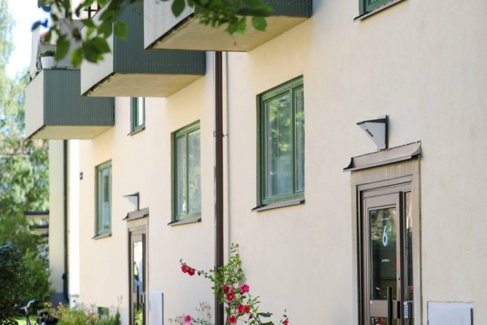 Entré till ett flerbostadshus med grönmålade balkonger och fönster, omgiven av grönska, blommande stockrosor och en parkerad cykel utanför dörren.
