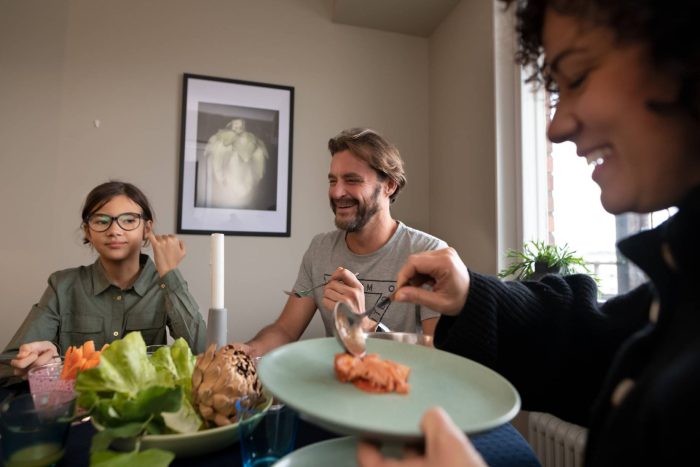 Tre personer sitter runt ett matbord och äter tillsammans. En vuxen serverar mat, en annan skrattar och ett barn tittar mot kameran. Maten består bland annat av grönsaker och lax. Bilden förmedlar en varm och vardaglig stämning i hemmiljö